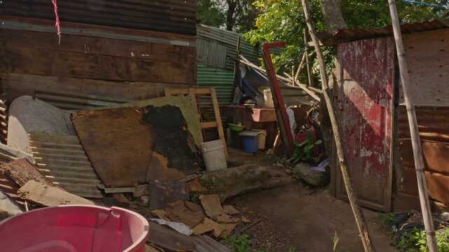 View of a narrow passageway or alley bordered by informal structures, wooden panels, and sheet metal, with various containers and jugs organized in the background.
