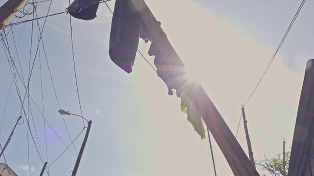 Low-angle shot looking up at a wooden utility pole and overhead wiring with hanging garments, as sunlight creates a strong lens flare.