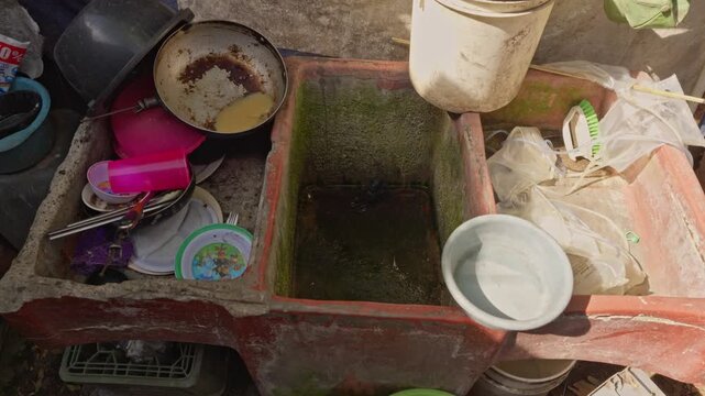 Close-up of concrete sink basins containing dirty dishes, a pan with food residue, and stagnant water with algae.