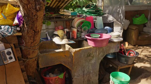 Wide panorama of an outdoor domestic space dominated by a concrete sink surrounded by kitchenware, a coiled green hose, and vegetation.