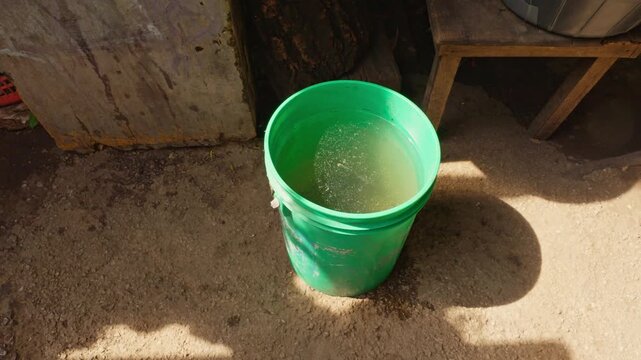 Cluster of assorted water containers, plastic jugs, and a bucket of cooking utensils near a makeshift wall structure.