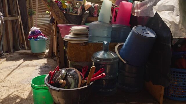 Perspective of a domestic storage and organization area with water jugs, buckets, and utensils clustered near the community pathway.