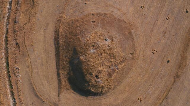 Cinematic top-down aerial drone shot of an ancient tumulus (mound) in a dry harvested field, tilting up to reveal a vast steppe landscape with hay bales.