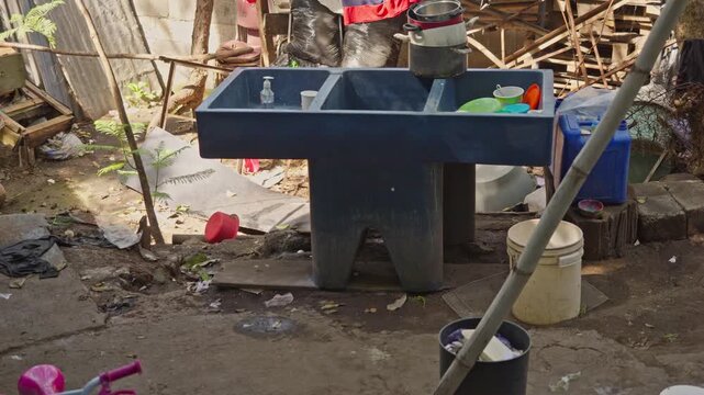 View of a blue plastic double-basin utility sink installed in a community yard, surrounded by everyday items and scattered vegetation.