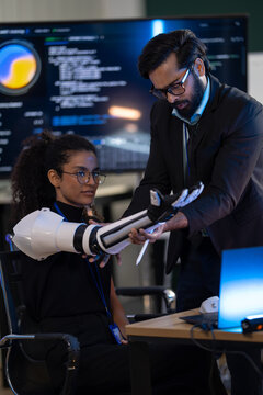 An engineer tests the articulated joints of a white prosthetic robot arm on the desk in a dark technology lab, during a productive late shift in the dark office.