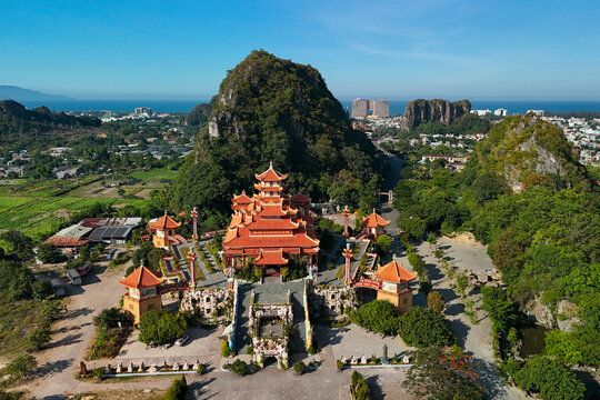 Traditional temple architecture sits surrounded by lush green landscapes and tall limestone cliffs overlooking a coastal city landscape on a clear day