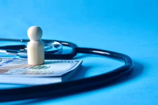 A wooden human figurine standing on a stack of US dollar banknotes next to a black medical stethoscope representing high healthcare costs.