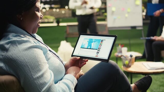 Black businessperson holds tablet with online concert video on streaming platform, music performance broadcast in work break, outdoor informal workspace, for mobile project planning.