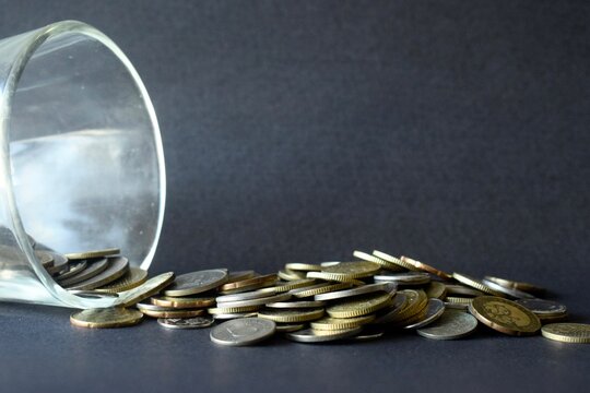 A close-up horizontal stock shot of a clear glass lying on its side with multiple coins flowing out from the opening onto a black background.