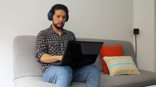 A caucasic latin man using a headset sitting on a grey couch working from home on a laptop
