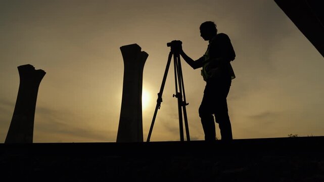 Silhouette of a survey engineer using surveying equipment at sunset with bridge columns in the background, representing infrastructure development and engineering precision.
