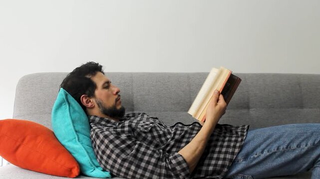 A caucasic latin man lying on a couch in a relaxing attitude leafing a text book