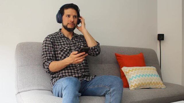 A latin man with beard sitting on a grey couch listening music on a headset and a smartphone in a happy attitude