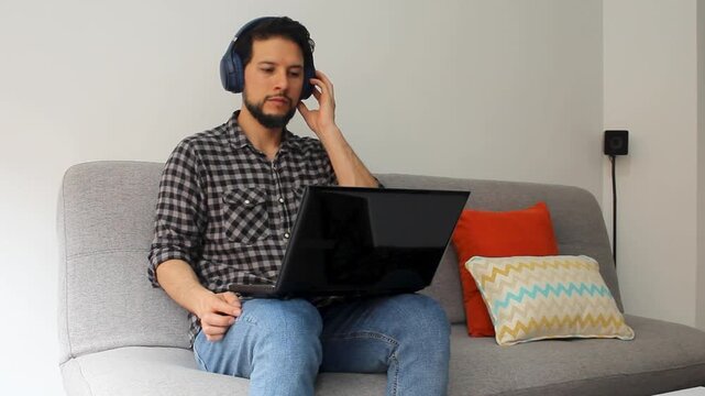 A latin man sitting on a grey couch listening music on a laptop over his legs with a headcase