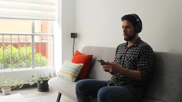 A Latin male at the right side of the screen listen music on a smartphone with a headset sitting on a living room