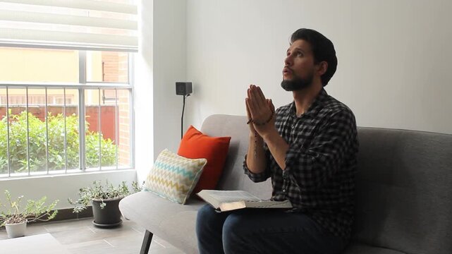 A latin man with a rosary bead on his hands and a bible on his legs sitting on a couch into a living room praying with the eyes on the sky