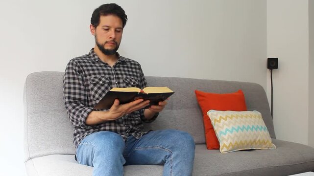 A latin young man sitting on a grey couch reading the holy bible in a concentrating attitude