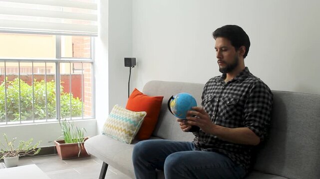 A latin man sitting on a grey couch spinning a small world globle in concentrating attitude