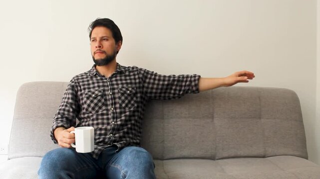 A relaxed hispanic male sitting at the left side of a couch drinking a beverage
