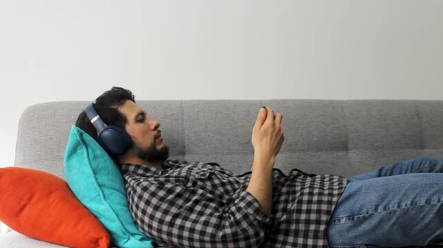 A latin caucasic man with beard lying on a couch watching something on a smartphone with a wireless headset in a concentrating attitude
