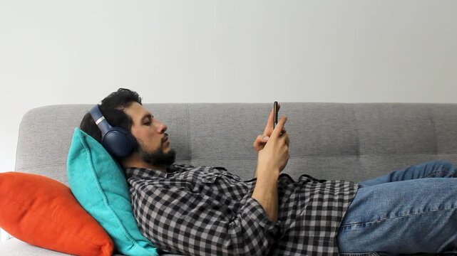 A latin man with beard lying on a couch typing on his smartphone with a wireless headset in a concentrating attitude
