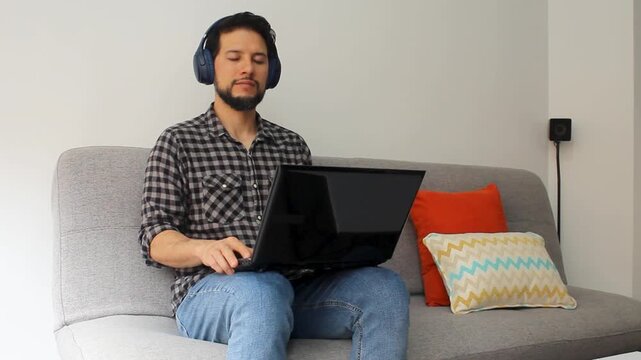 detailed view of a latin man with his eyes closed sitting on a couch listening music on a wireless headset and a laptop over his legs in a happy attitude