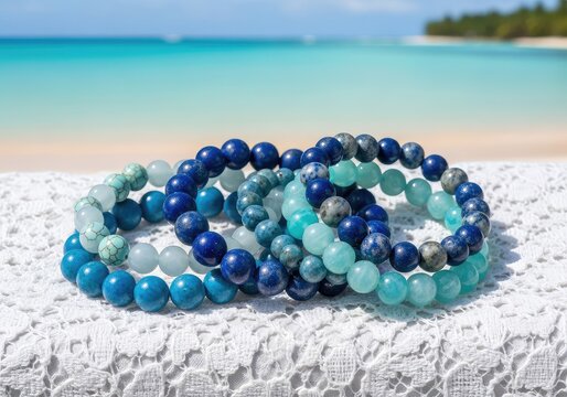 Close up of handmade natural blue stone bracelets on a white lace fabric with a blurred turquoise ocean background and sandy tropical beach, coastal, resort, lapis