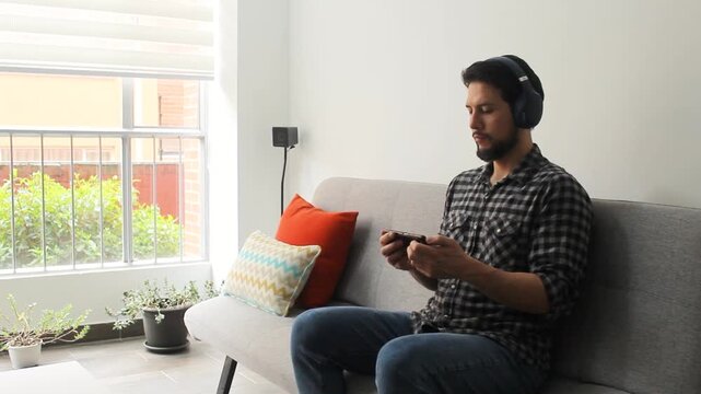 A caucasic male sitting at the right side of a couch listening and watching a video music in a happy attitude