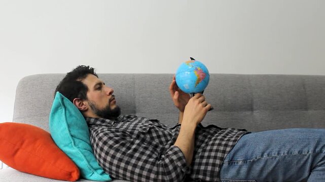 A latin caucasic man with beard lying on a couch watching and spinning a small world globle in concentrating attitude