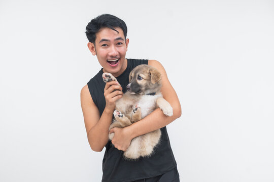 A cheerful young Southeast Asian man carries a cute puppy and playfully makes it wave, capturing a joyful pet-owner moment.