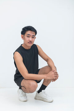 A young filipino male in athletic clothing squats comfortably while looking at the camera in a clean studio setting.