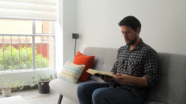 A latin male sitting on a grey couch in the morning reading the holy bible in a peaceful attitude