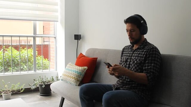 A caucasic hispanic man sitting on a grey couch texting with his phone and using a headset in a serious attitude