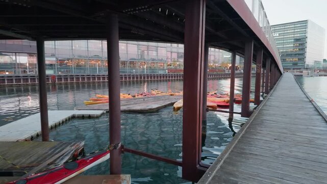 Wooden dock canoe bridge city, gimbal stabilizer shot. 60fps wide push in walking forward on pier looking at red orange kayaks under metal overpass in Copenhagen.