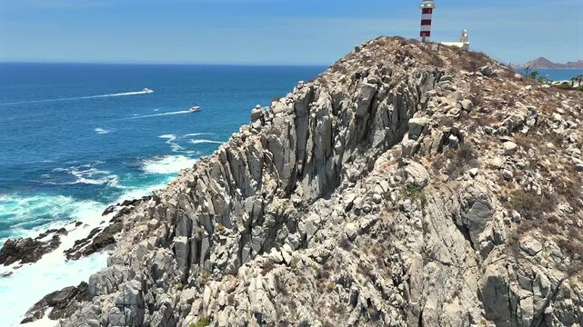 Drone climbs and orbits the Cabo San Lucas cliffside to reveal El Encanto Lighthouse and a church above, with yachts crossing the water and the Arch of Cabo San Lucas visible in the far distance.
