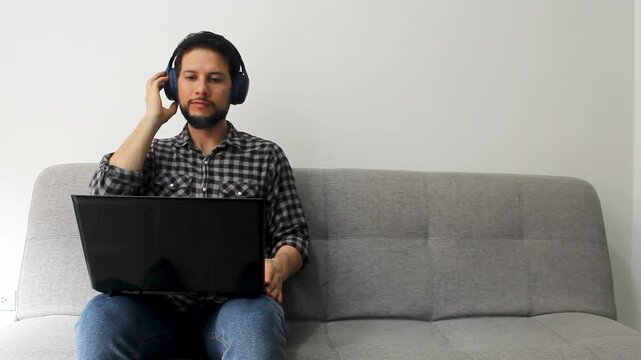 Detailed view of a smiling male sitting at the left side of a grey couch listen music on a wireless headset with a laptop over his legs