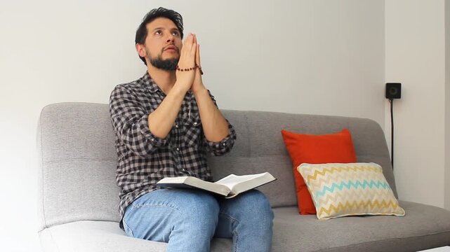 Detailed view of a caucasic man sitting at the left side of a couch praying with a rosary on his hands and a holy bible over his legs and looking to the sky
