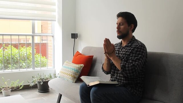 A latin man sitting on a couch praying with a rosary and a holy bible over his legs