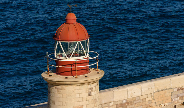 Primer plano de la c&uacute;pula roja de un faro de piedra tradicional junto a las profundas aguas azules del mar Mediterr&aacute;neo en un d&iacute;a soleado.