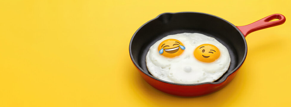 Fried egg with laughing emoji faces in a red frying pan on a yellow background. Creative breakfast for World Emoji Day. Funny cuisine concept for social media content and humor. Banner with copy space
