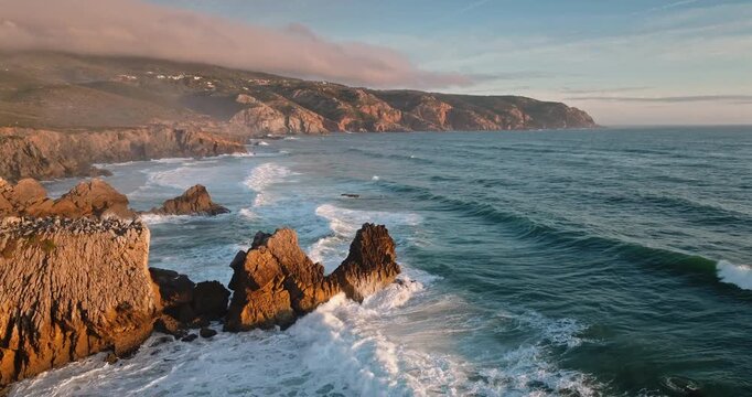 Rugged coastline of Sintra Cascais Natural Park with dramatic ocean waves crashing against rocks and towering cliffs with golden light during a beautiful sunset. Travel Portugal. Aerial drone panorama