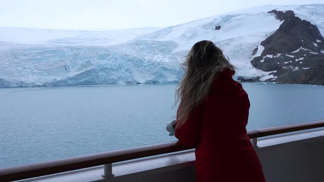 Woman Photographer on Antarctica Cruise Ship Stands with Professional Camera in Orange Red Coat. Wind Blows Hair. Snow Solo Female Travel Beautiful