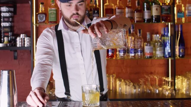 Cold beverage. Cocktail glass. Skillful male bartender straining liquid using sieve with ice cubes at bar counter.