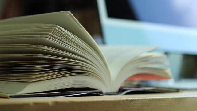 Horizontal 4K side angle video of an open book on a wooden desk with pages fluttering gently and a blurred desktop monitor behind