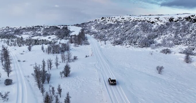 Drone view of a 4x4 driving along a snowy Hei&eth;m&ouml;rk road in Iceland, with parallel tire tracks, winter hills, bare trees, and open frozen terrain.