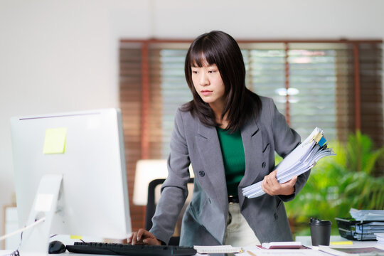 Asian businesswoman multitasking at office desk, managing documents and using computer, representing professional productivity, diligent work, and corporate efficiency