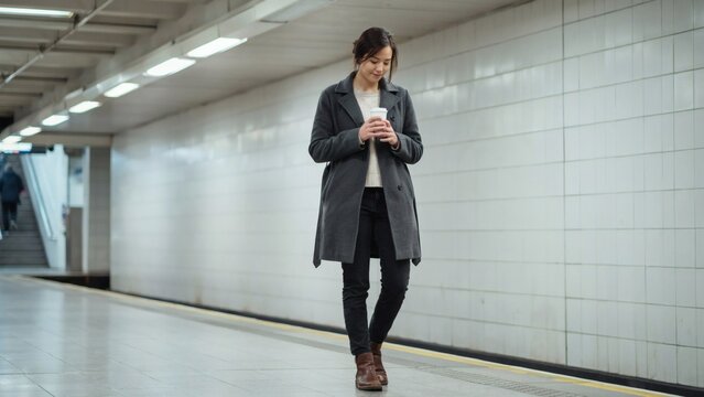 Young woman walking along an empty subway station platform while holding a warm beverage during her daily commute