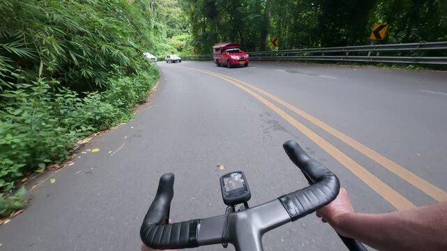 First-person POV footage of a road cyclist descending a winding mountain road surrounded by lush tropical forest. Passing vehicles on a scenic curve add a realistic transportation atmosphere, making t