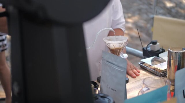 A man carefully pouring hot water from a long-spout kettle through a coffee filter during a pour-over brewing process at a Thai outdoor market. Coffee brewing concept