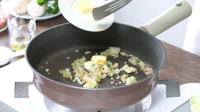 Close-up of butter being added to a frying pan with saut&eacute;ed chopped onion and garlic. A home cook prepares the aromatic base for a delicious meal in a modern kitchen. Healthy homemade cooking, recipe 